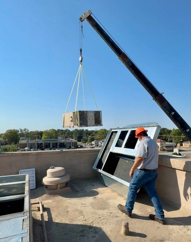 A construction worker in a hard hat guides a crane-lifted air conditioning unit onto a rooftop.