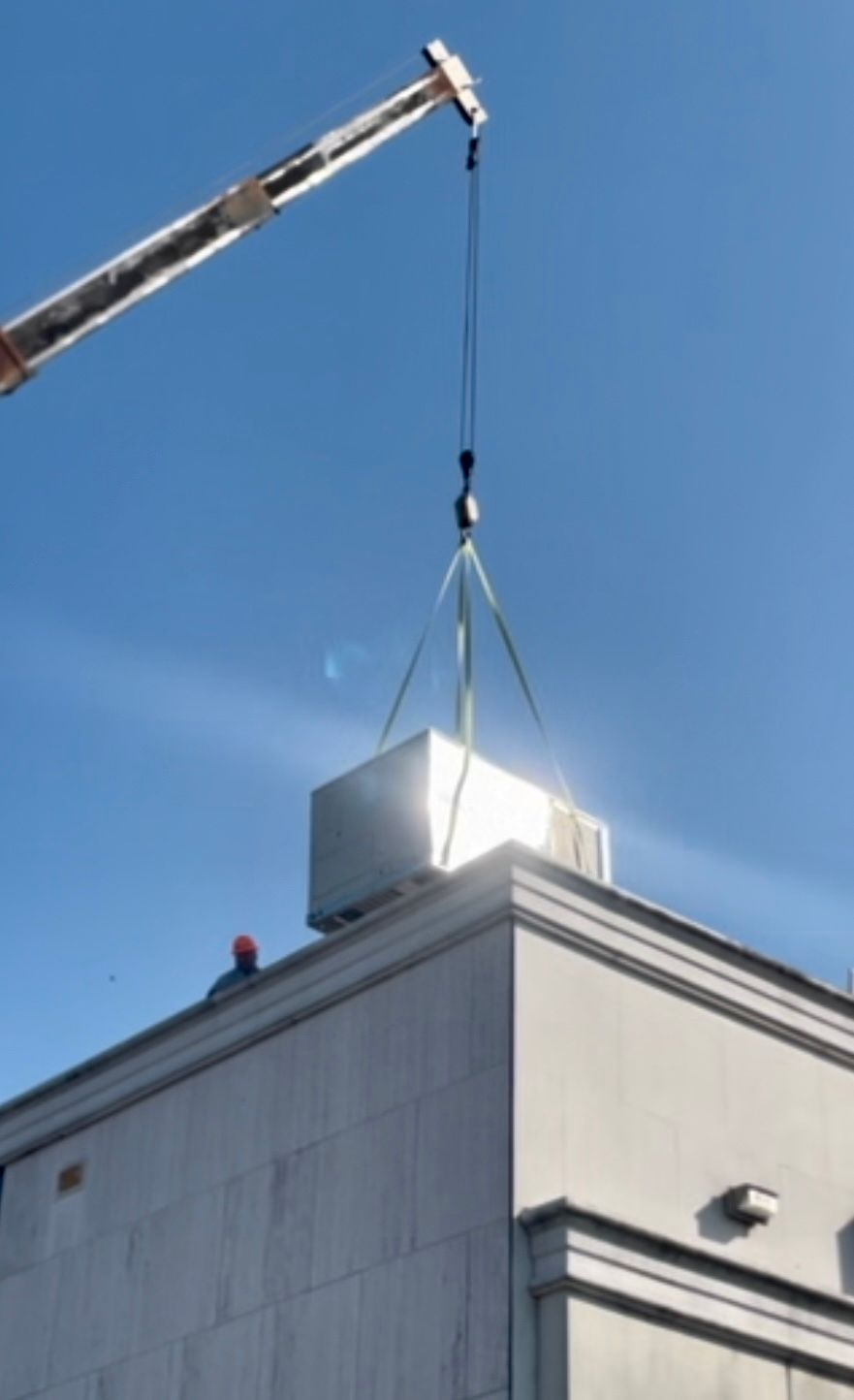 A construction crane lowers a large metal HVAC unit onto the roof of a building under a clear blue sky.