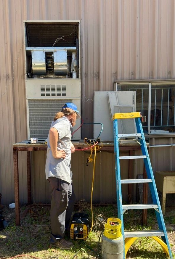 A technician stands beside an outdoor HVAC unit on a metal stand, working with tools and a refrigerant tank.
