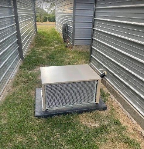 An outdoor air conditioning unit sits on a black pad between two gray, corrugated metal storage buildings.
