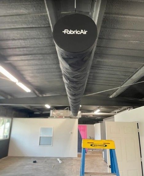 A black fabric air duct hanging from the ceiling of an unfinished room with a ladder in the foreground.