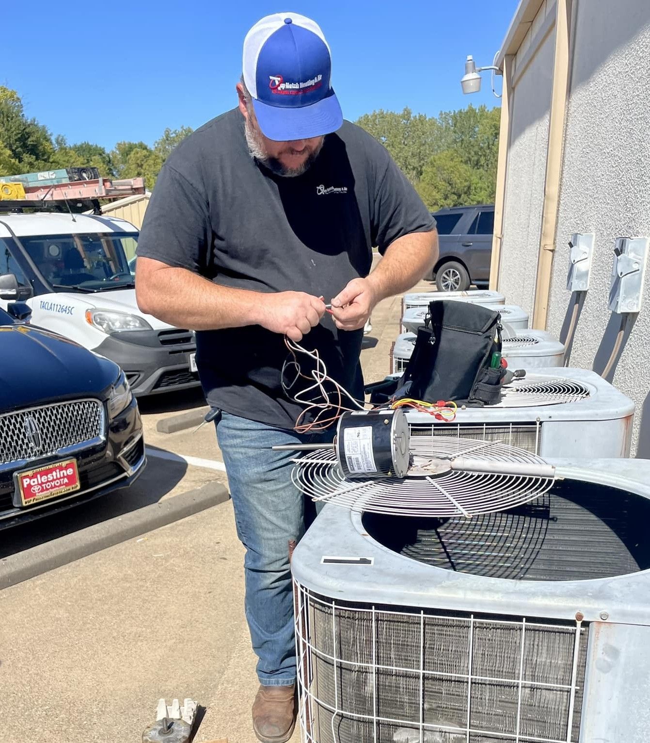 A person wearing a blue and white cap works on the wiring of an outdoor HVAC unit in a parking lot.