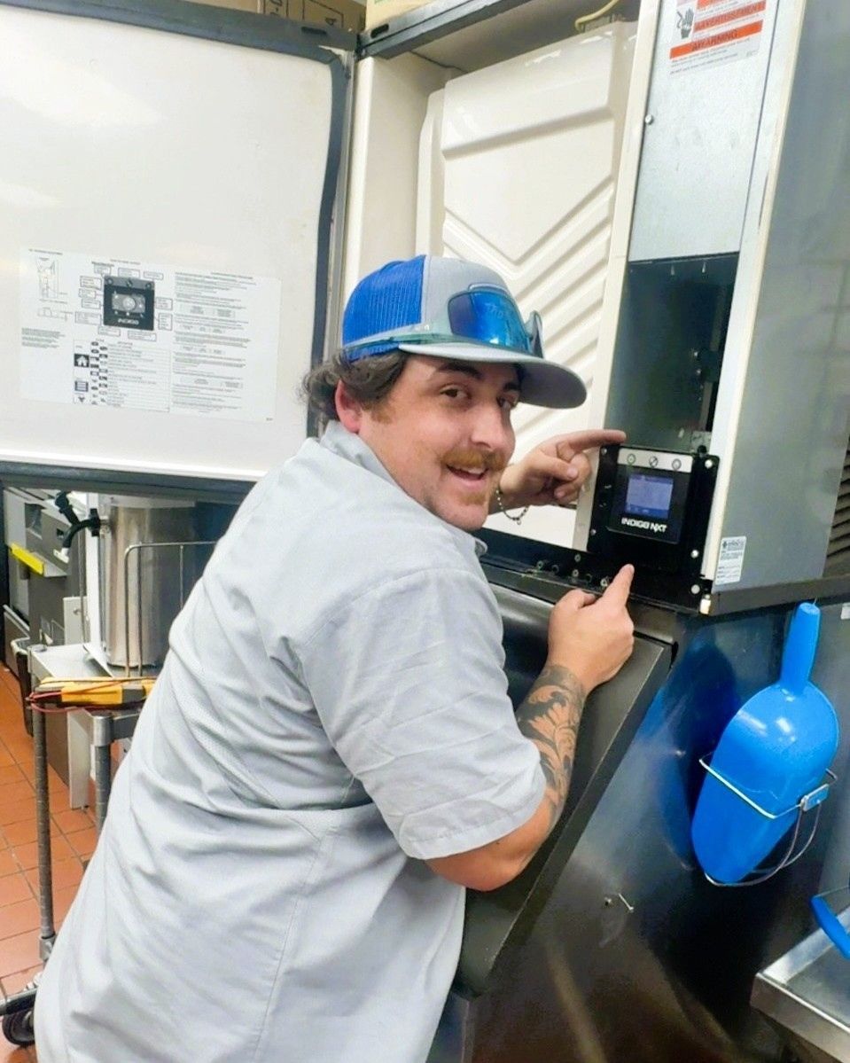A person with a mustache and cap points to a control panel on a commercial ice machine in a kitchen setting.