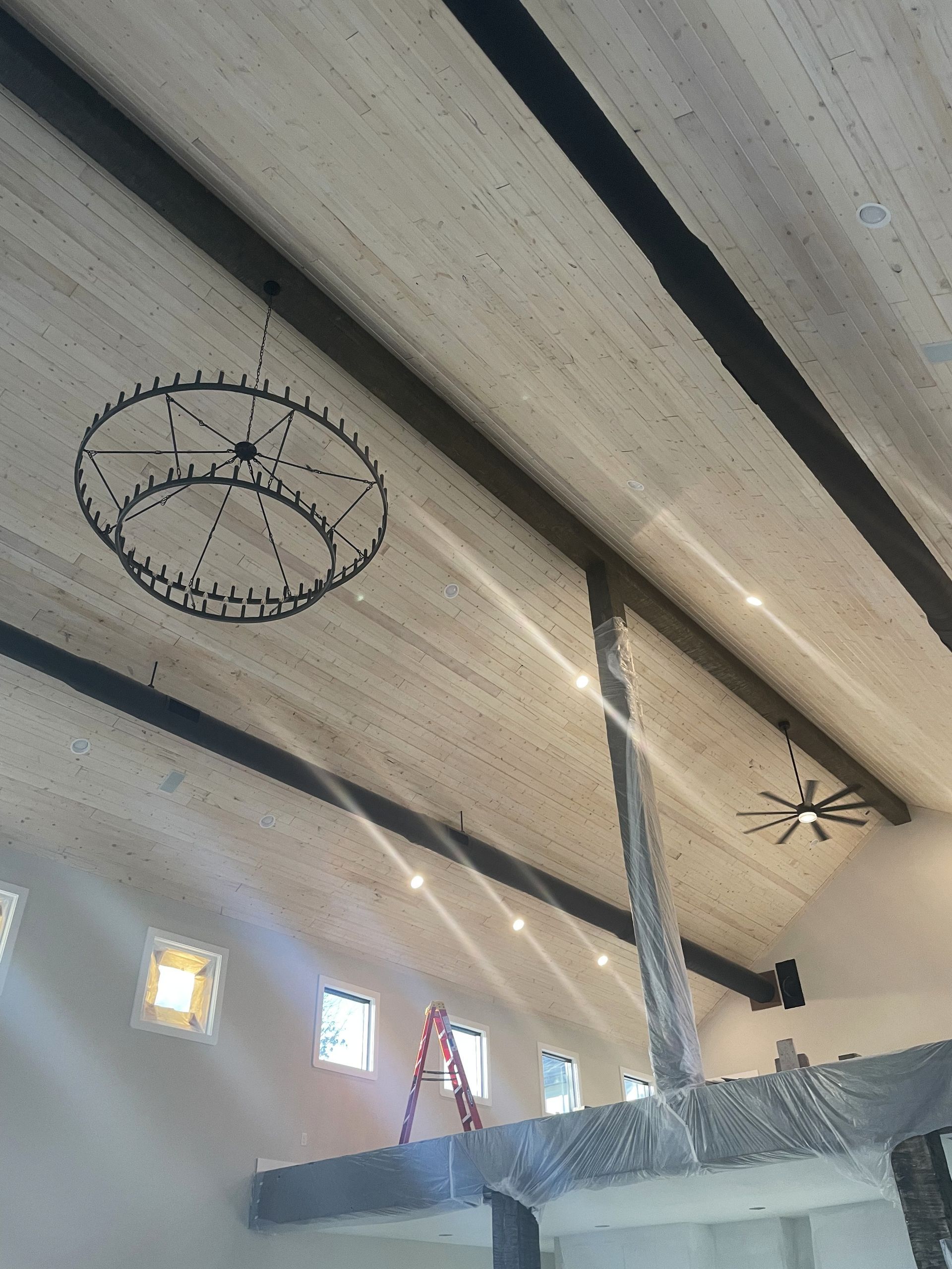 A low-angle view of a room with a high, vaulted wooden ceiling featuring exposed dark beams, a round chandelier, and a fan.