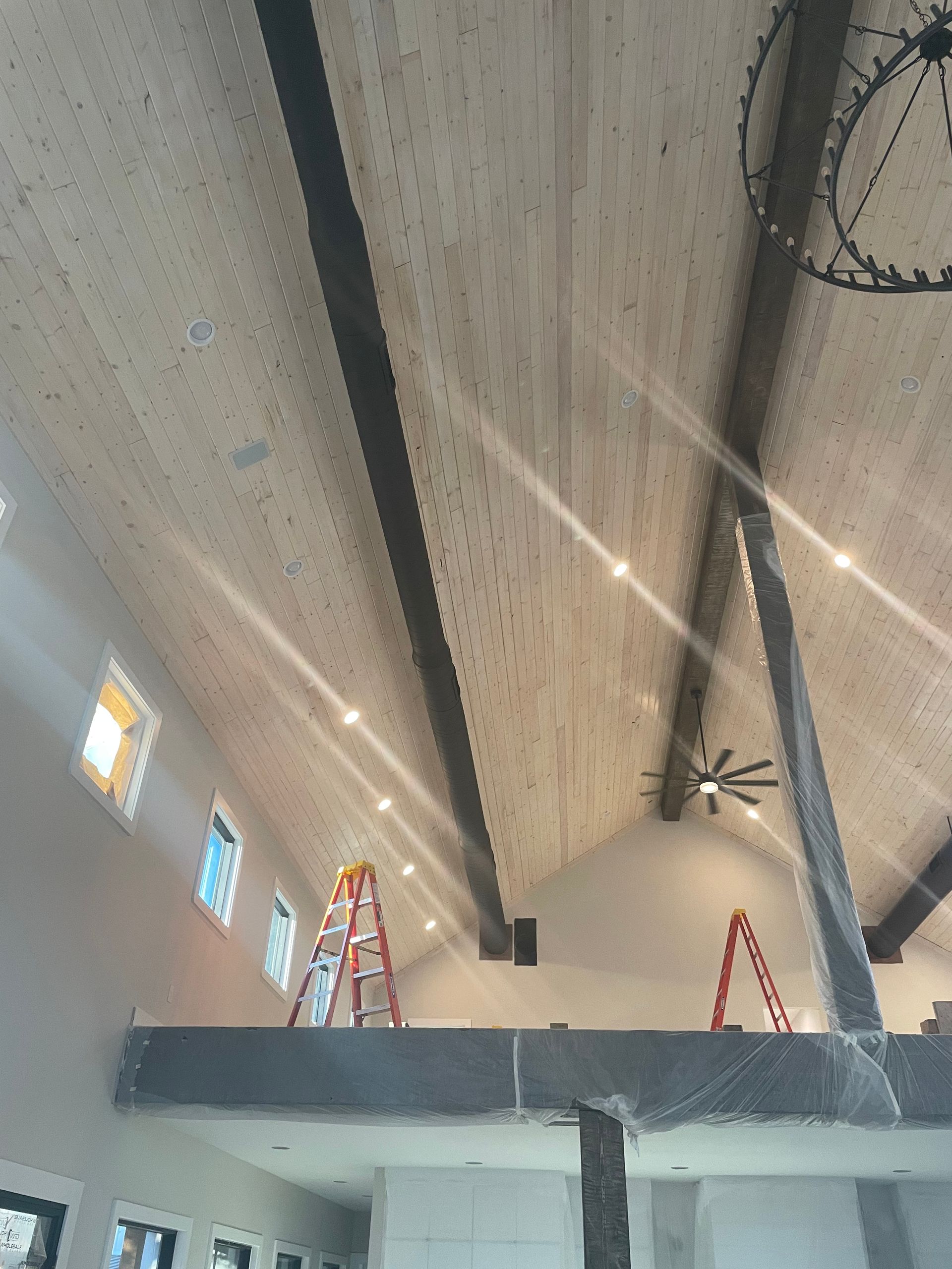 An interior view of a room under construction with wood-paneled vaulted ceilings, dark beams, and two A-frame ladders.