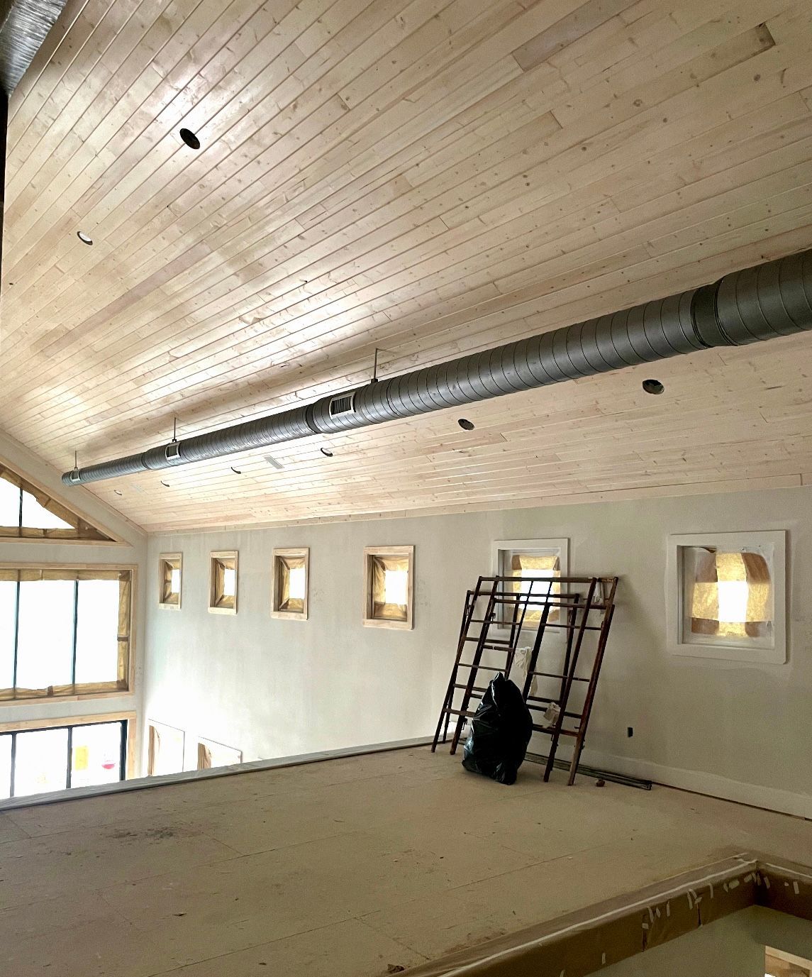 An interior view of a room under construction featuring a vaulted wood-paneled ceiling, exposed ductwork, and wall windows.