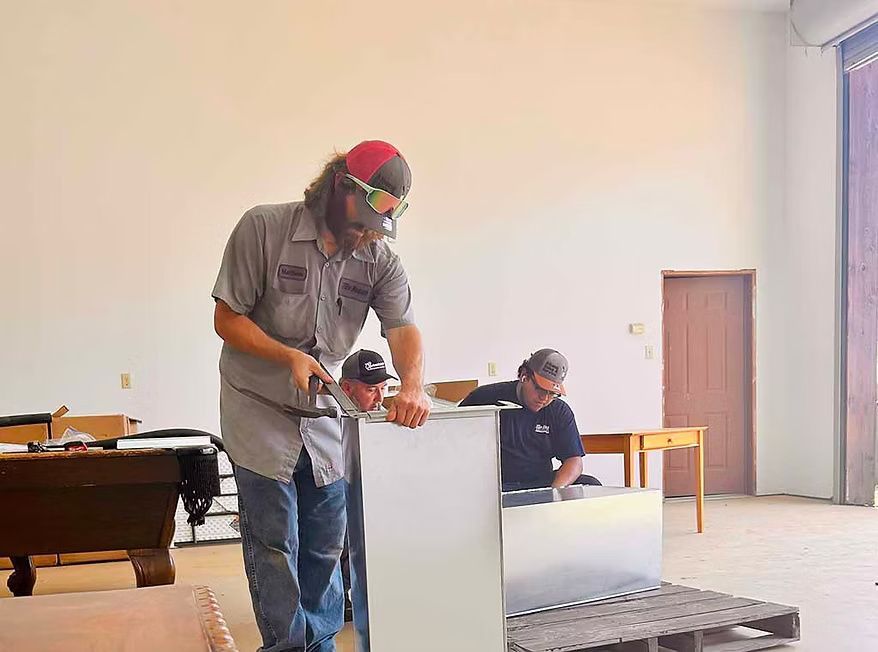 Three people working with metal in a bright workshop; one is using a tool on a large white metal object. Three people working with metal in a bright workshop; one is using a tool on a large white metal object.