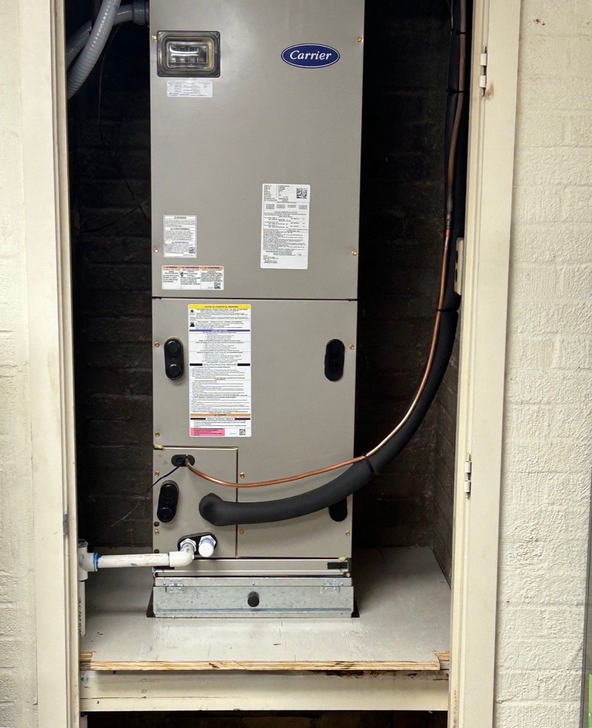 A tan Carrier HVAC unit sits inside a small, recessed closet against a brick wall.