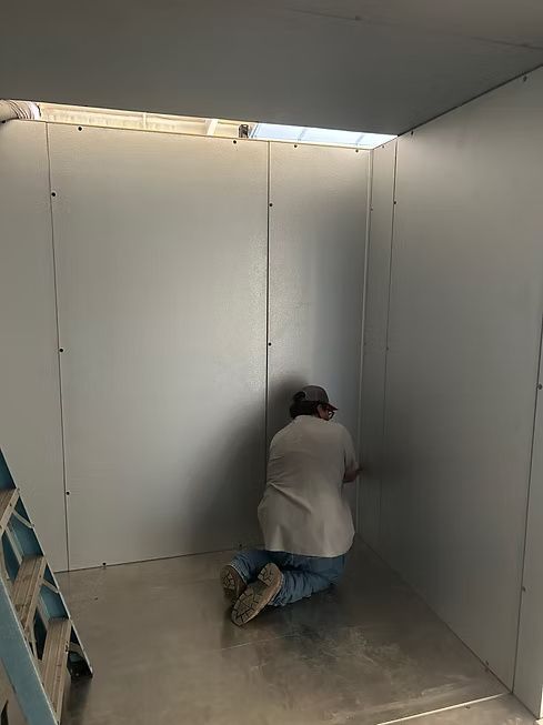 Person kneeling in a metal-walled room, working on the corner. A stepladder is visible on the left. Person kneeling in a metal-walled room, working on the corner. A stepladder is visible on the left.