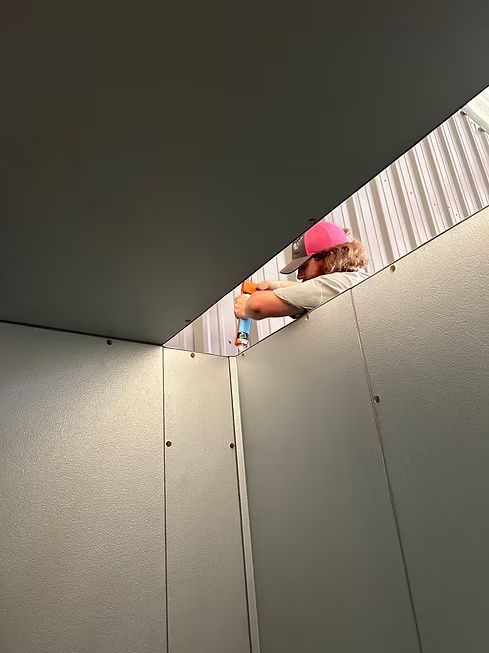 Person in a pink hat working inside a structure with gray walls, holding a tool near the ceiling. Person in a pink hat working inside a structure with gray walls, holding a tool near the ceiling.