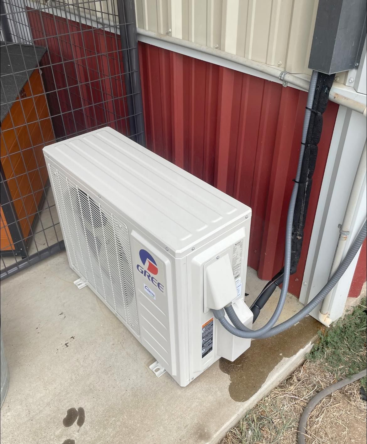 A white Gree air conditioning outdoor unit installed on a concrete pad against a red metal wall.