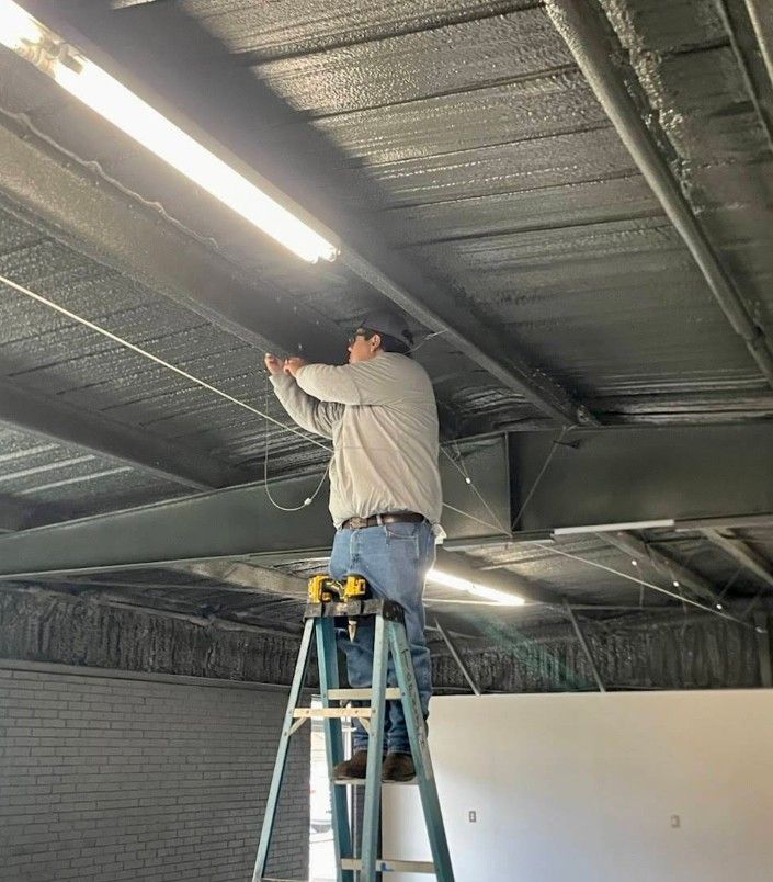 A person in a light-colored shirt and jeans stands on a ladder working on ceiling lights in an industrial space.