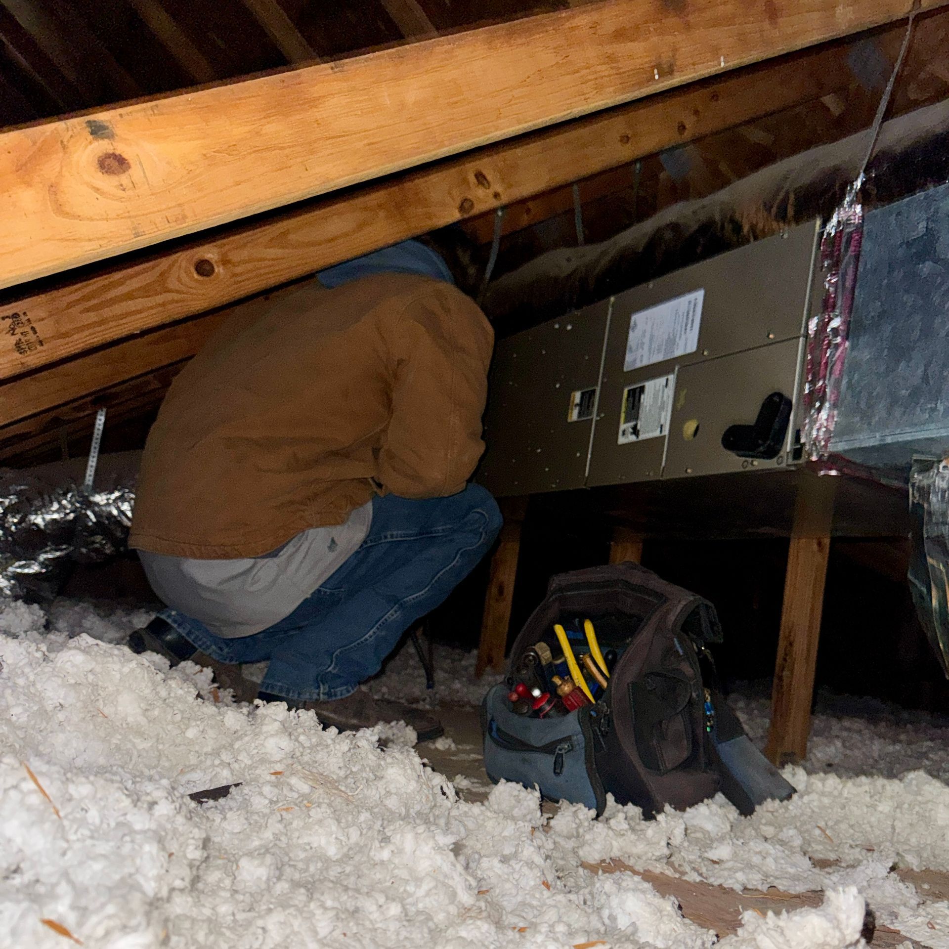 A technician wearing a tan jacket works on an HVAC unit in a dark, insulation-filled attic with a tool bag nearby.