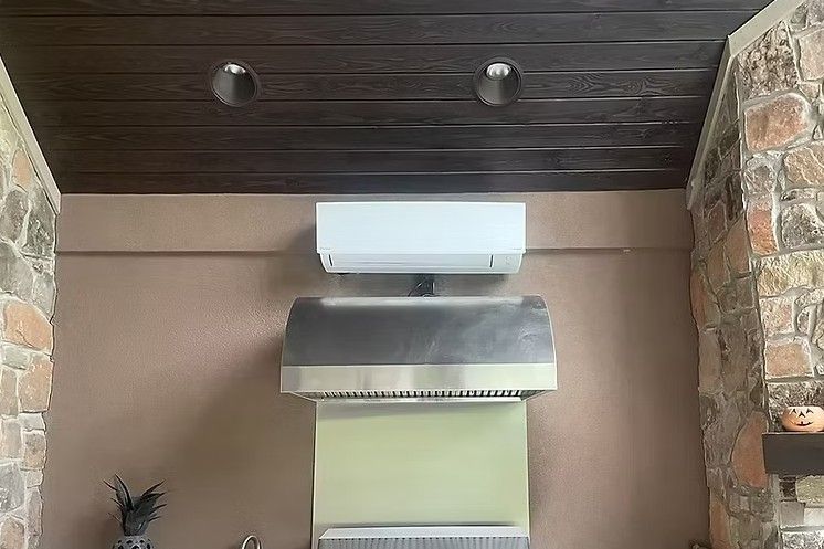 A white mini-split air conditioner is mounted on a brown wall above a stainless steel range hood in a stone-walled room.