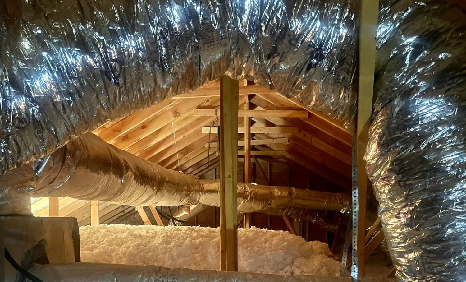 Attic space featuring wooden rafters, thick fiberglass insulation, and silver-colored flexible air ducts.