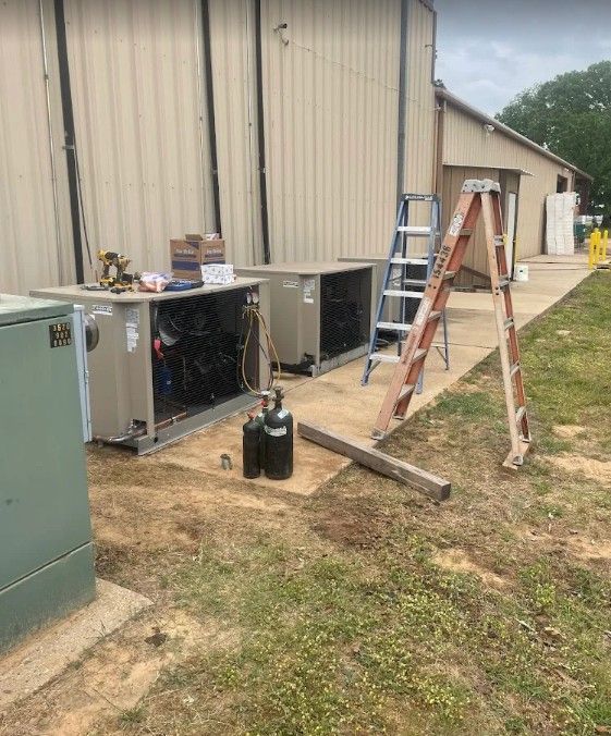 Two HVAC units sit against a beige metal building with a ladder and two gas cylinders on the concrete pad.