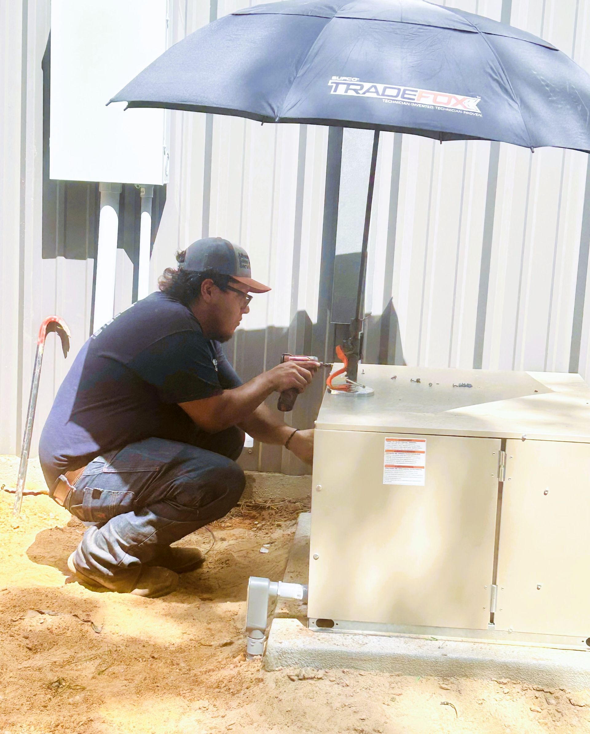 A technician in a baseball cap crouches outside to service an HVAC unit under a dark umbrella.