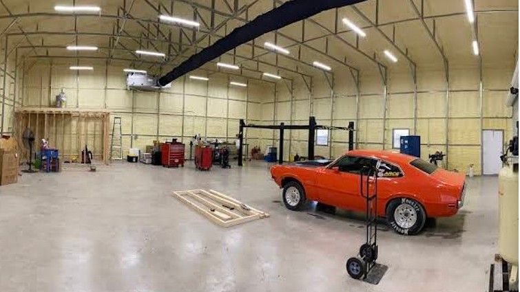 A bright, spacious workshop with insulated walls, a car lift, and a vibrant orange muscle car parked on a concrete floor.