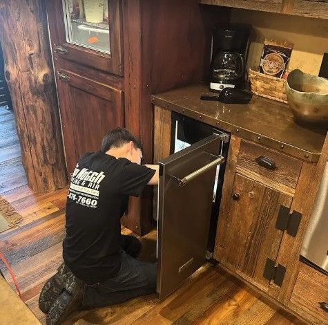A person wearing a black uniform shirt kneels on a wooden floor, repairing a small under-counter refrigerator.