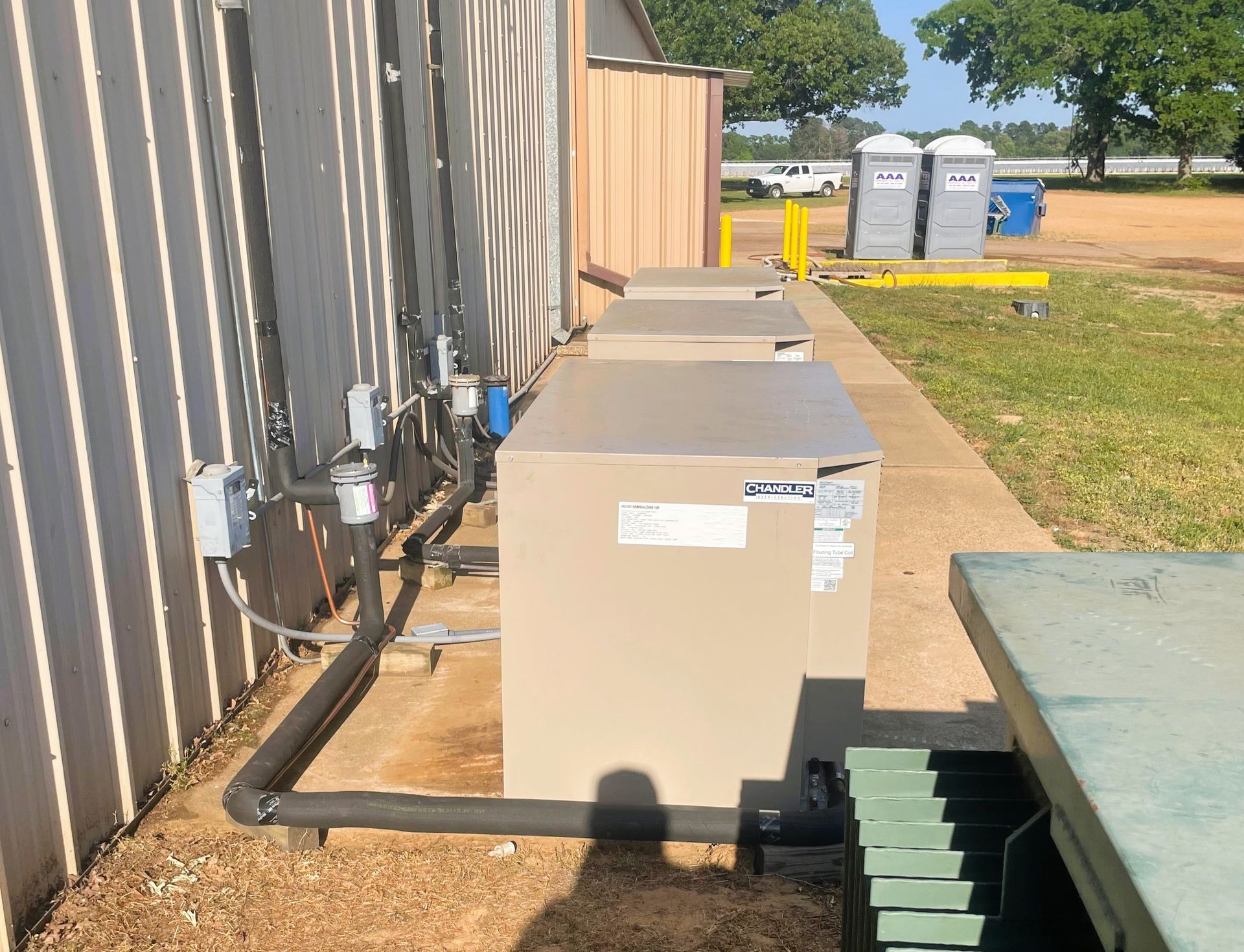 Three beige HVAC units sit on a concrete path along a metal-sided building, with blue portable toilets in the background.