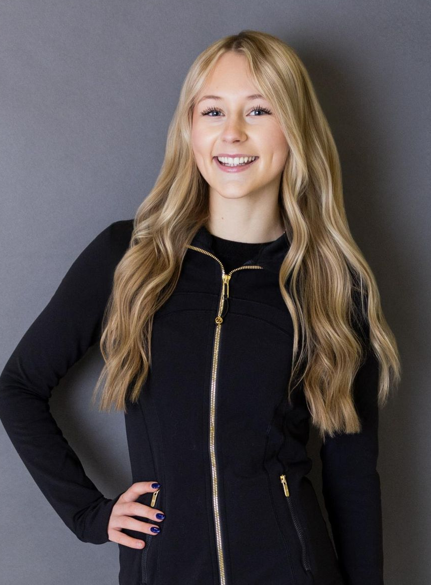 Woman smiles, standing on an ice rink, wearing a black vest and long sleeves.