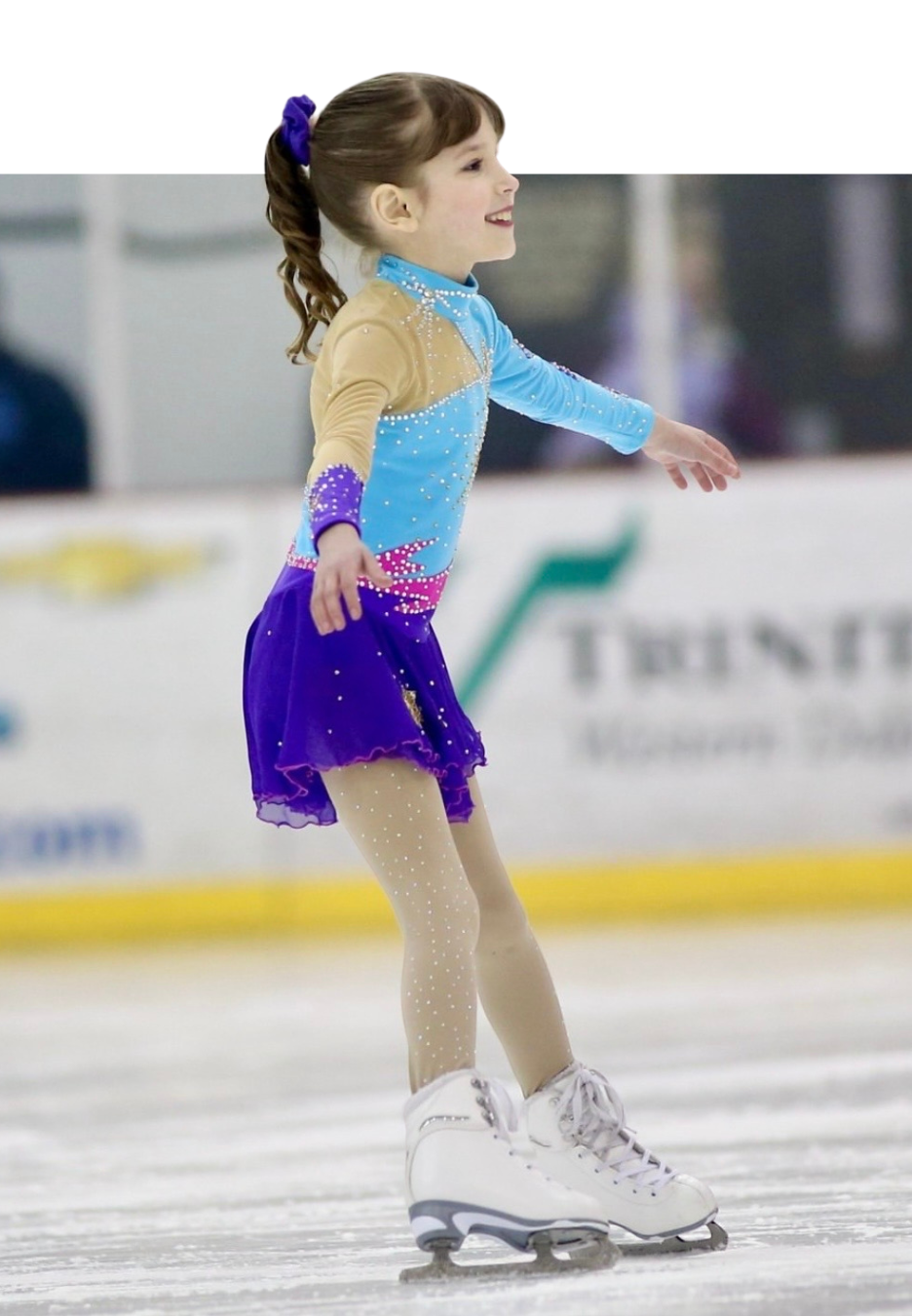 Young ice skater in a blue and purple costume performing on the ice, smiling.