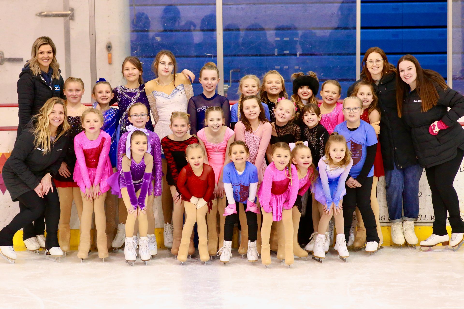 Group of figure skaters posing indoors in colorful costumes and black jackets on an ice rink