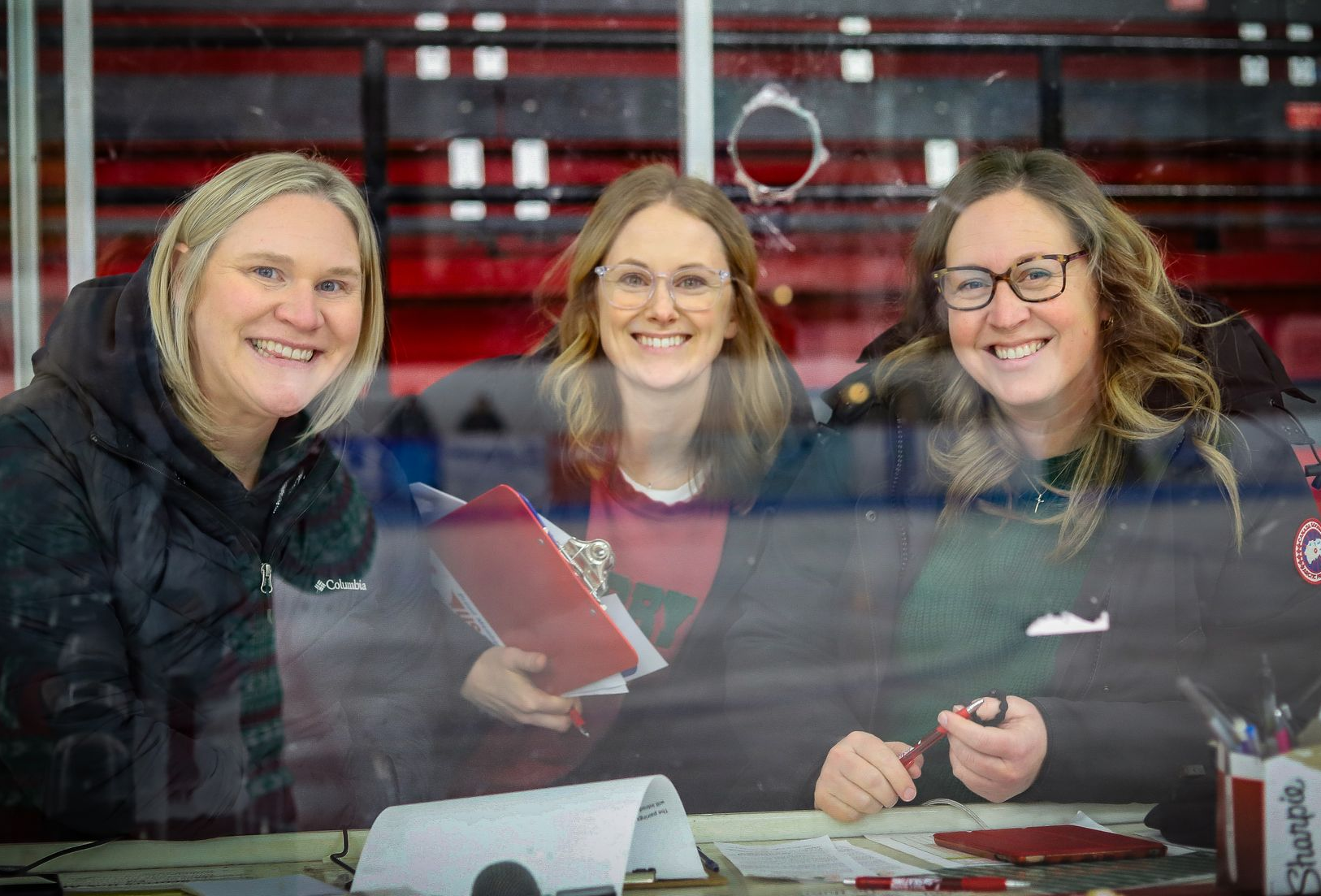 Three smiling people behind a table, possibly at an event, with red and white background.