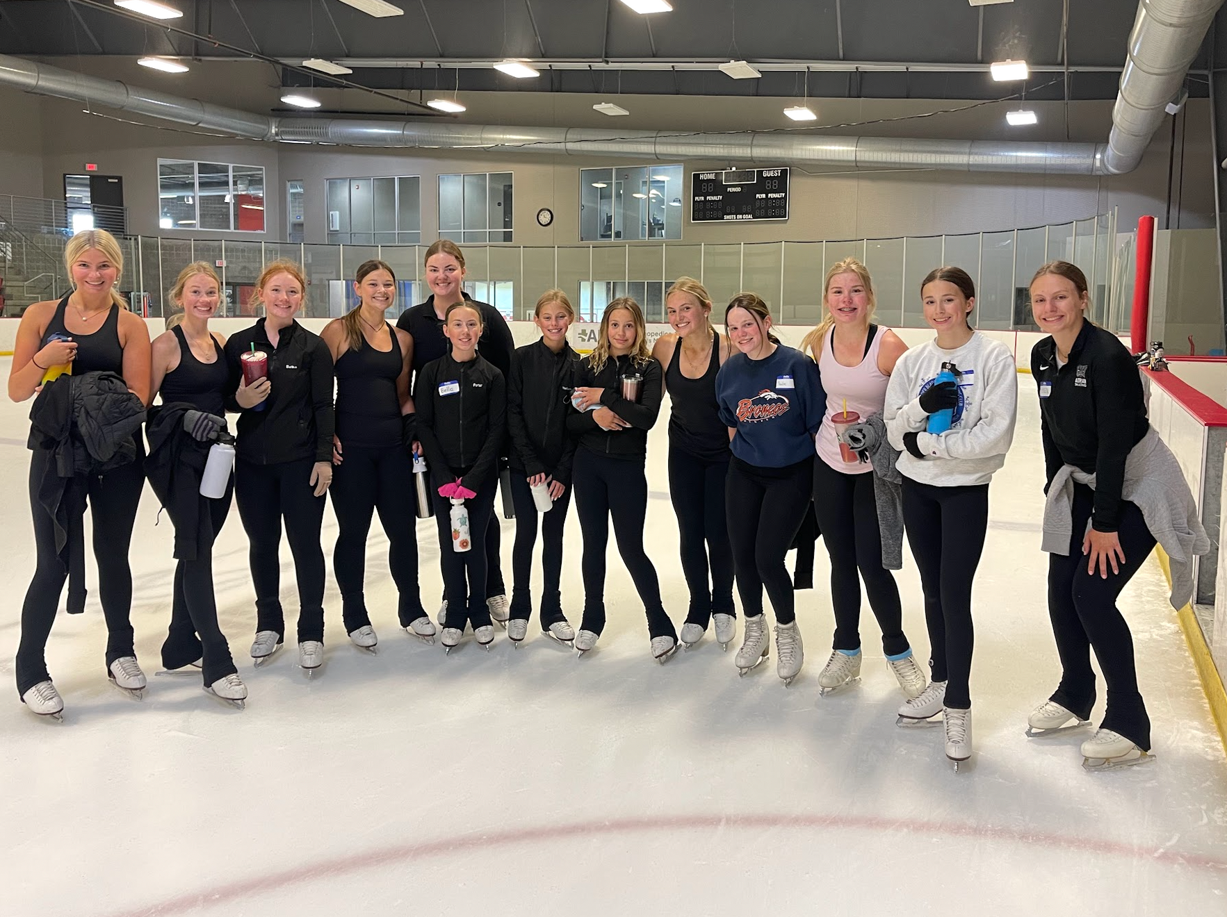 Group of skaters posing on an ice rink. They wear skates and dark athletic clothing, smiling. Red barrier visible.