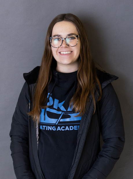 Woman with long blonde hair, smiling, wearing black jacket with gold zipper and black pants.