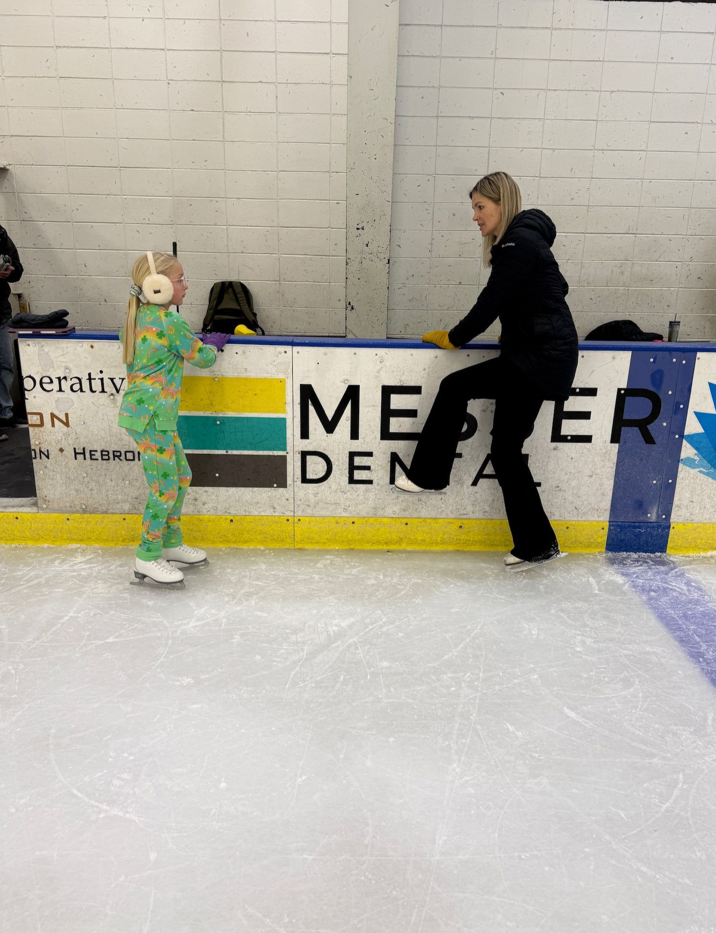 Two people on an indoor ice rink near the boards, one in black and one in green, with “MEER” signage behind them.