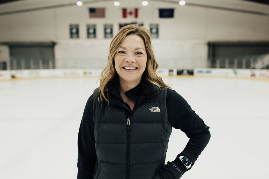 Woman smiles, standing on an ice rink, wearing a black vest and long sleeves.