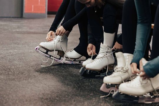 People in black pants tying white ice skates, seated on a bench, waiting to skate.