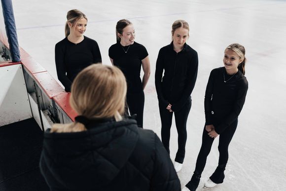 A woman in a black jacket talks to four ice skaters in black outfits on an ice rink.
