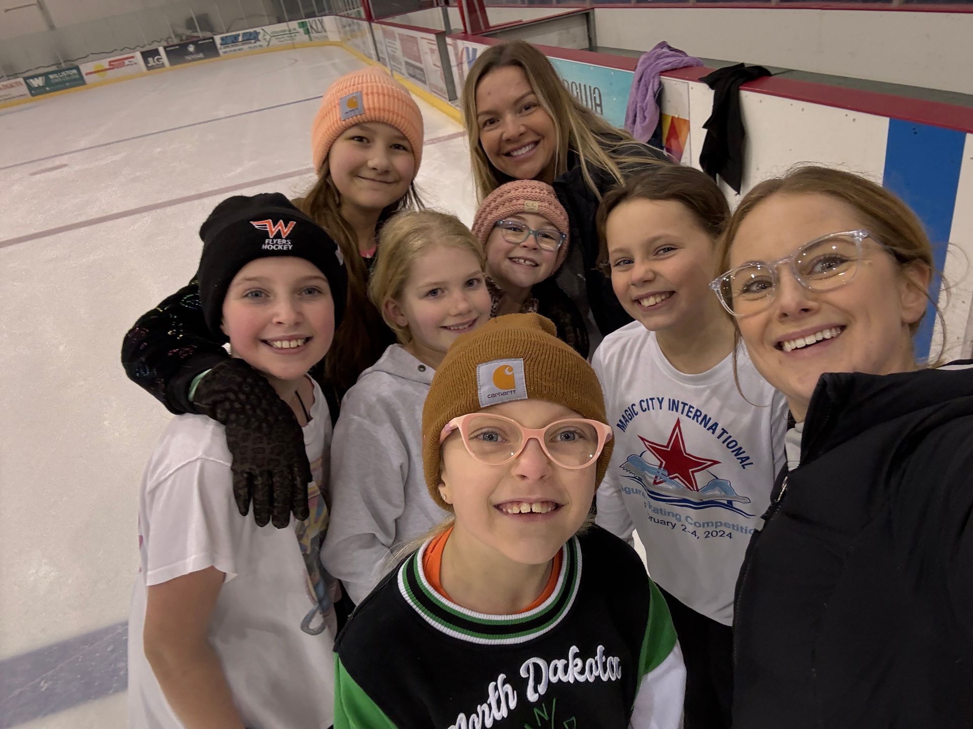 Five girls in ice skates, smiling and posing together on an ice rink.