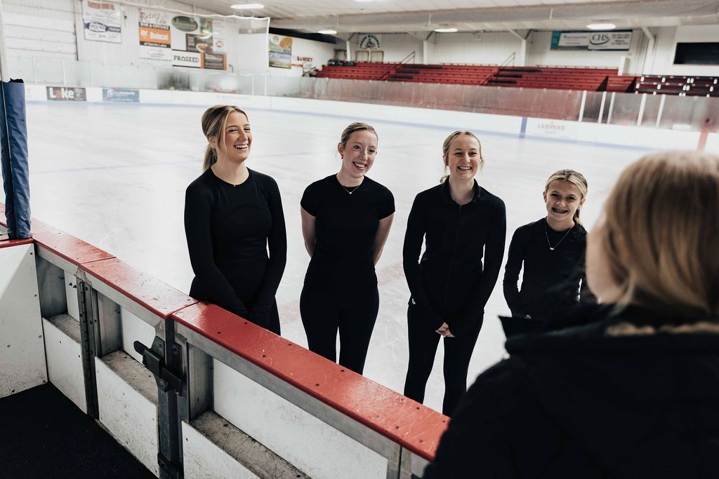 Four people on ice rink, looking at a person in the foreground. They are wearing black. Rink has red seating.
