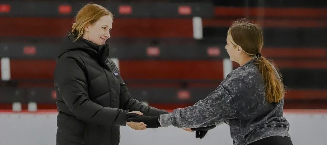 Two people ice skating, holding hands. One wears a black jacket, the other a gray sweatshirt.