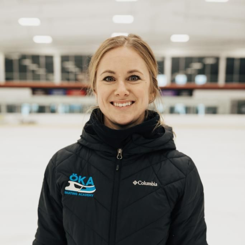 Woman smiling, wearing a black jacket, standing in an ice rink with an 