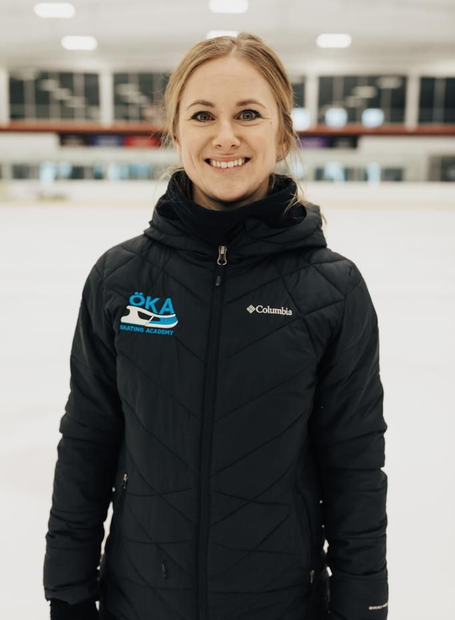 Woman smiles, hands on hips, wearing a black jacket; gray backdrop.