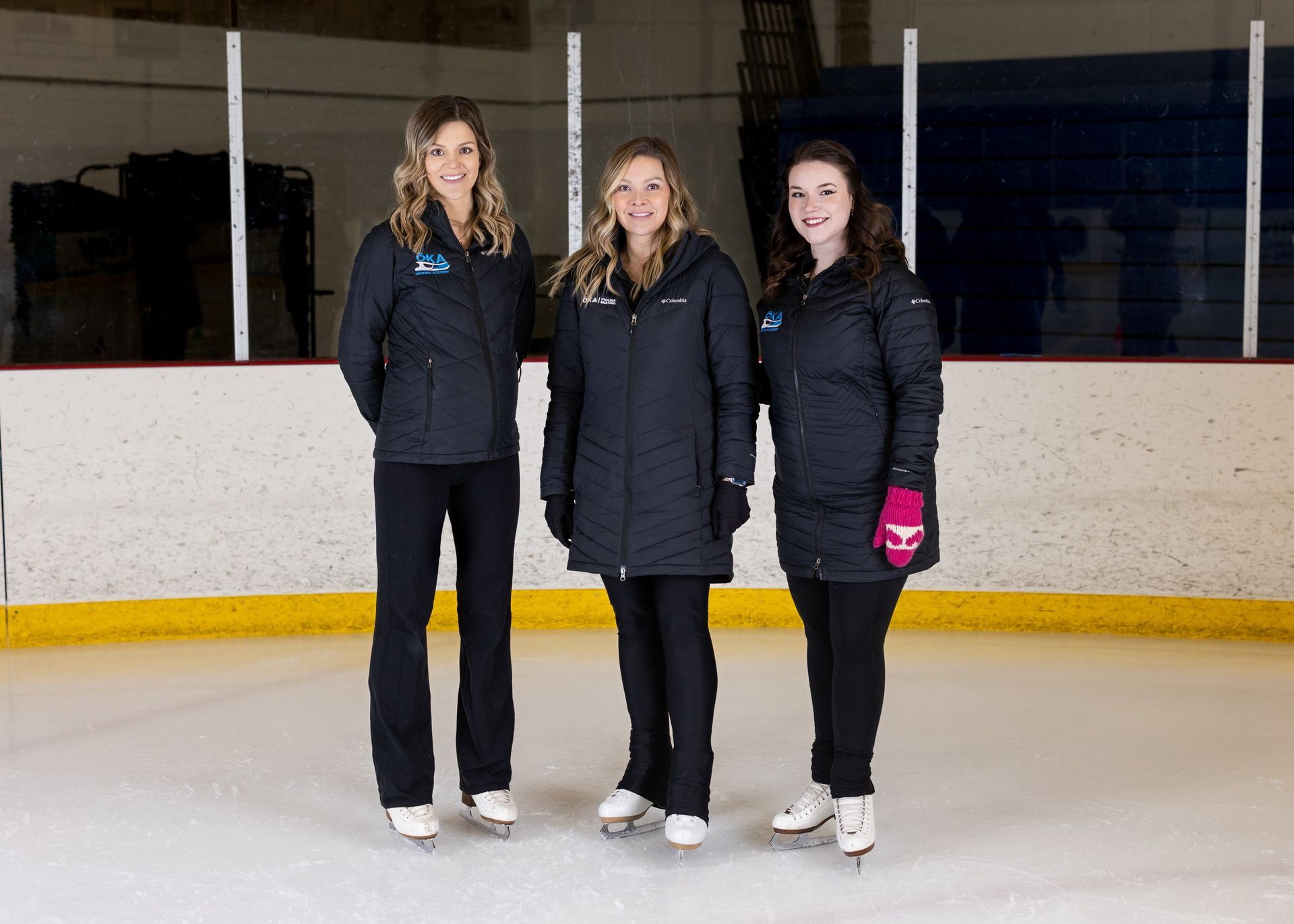 Three women in black jackets and pants stand on an ice rink, wearing ice skates.