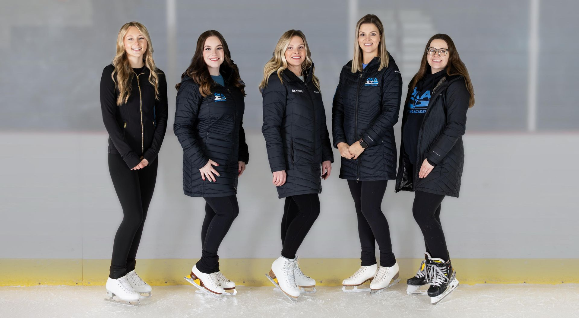 Five women in black coats and ice skates pose on an ice rink.