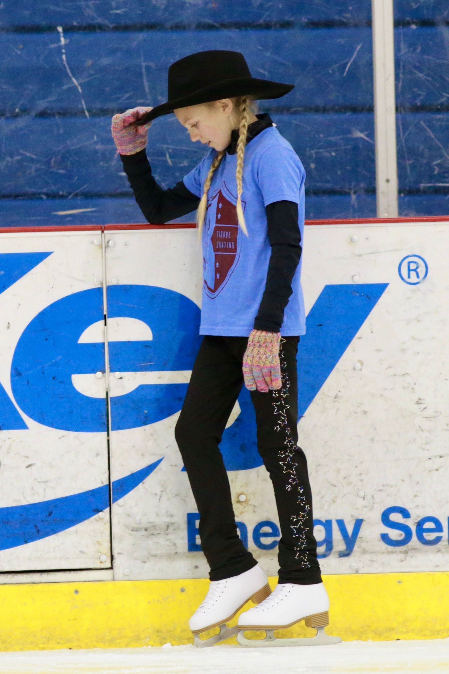 Skater in black hat and blue shirt standing on an indoor ice rink, looking down with hand on hat
