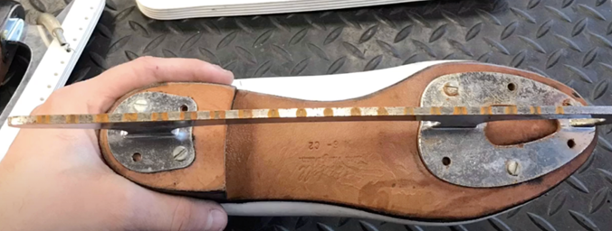 A close-up of a figure skate boot held by a hand. The blade is being measured.