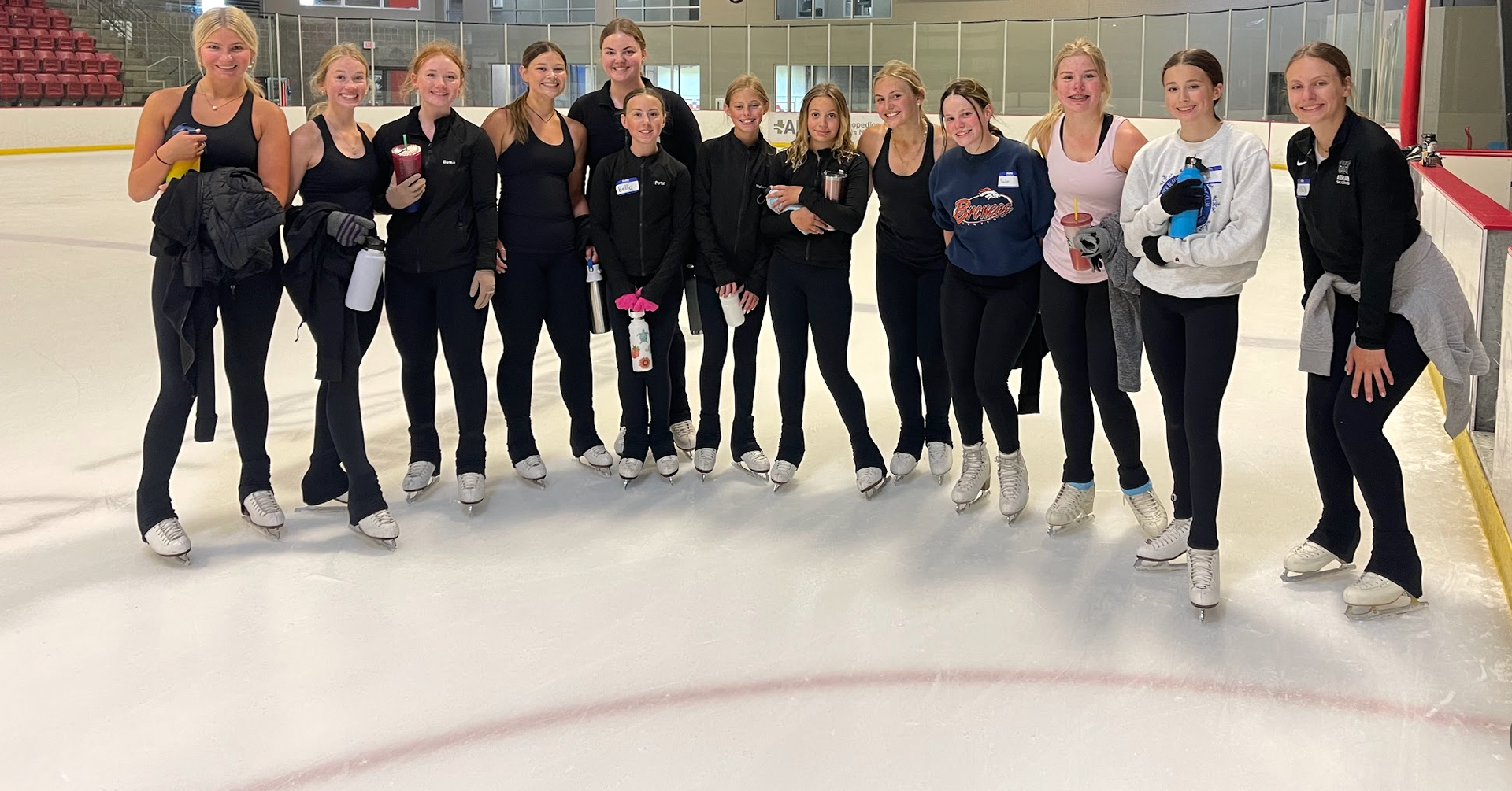 A group of children on an ice rink, many in skates, wearing matching shirts, smiling.