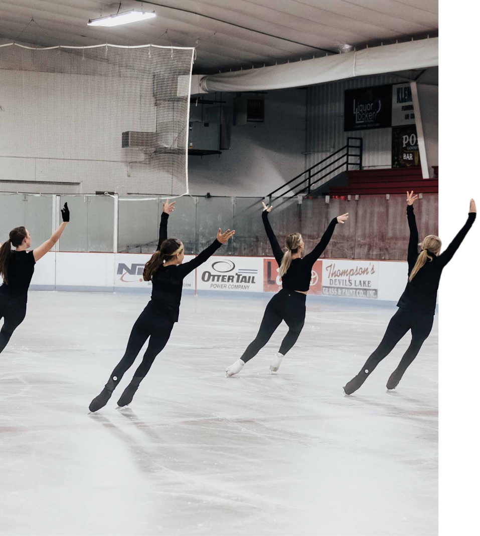 Four figure skaters in black outfits on ice, arms raised, skating together in an indoor rink.