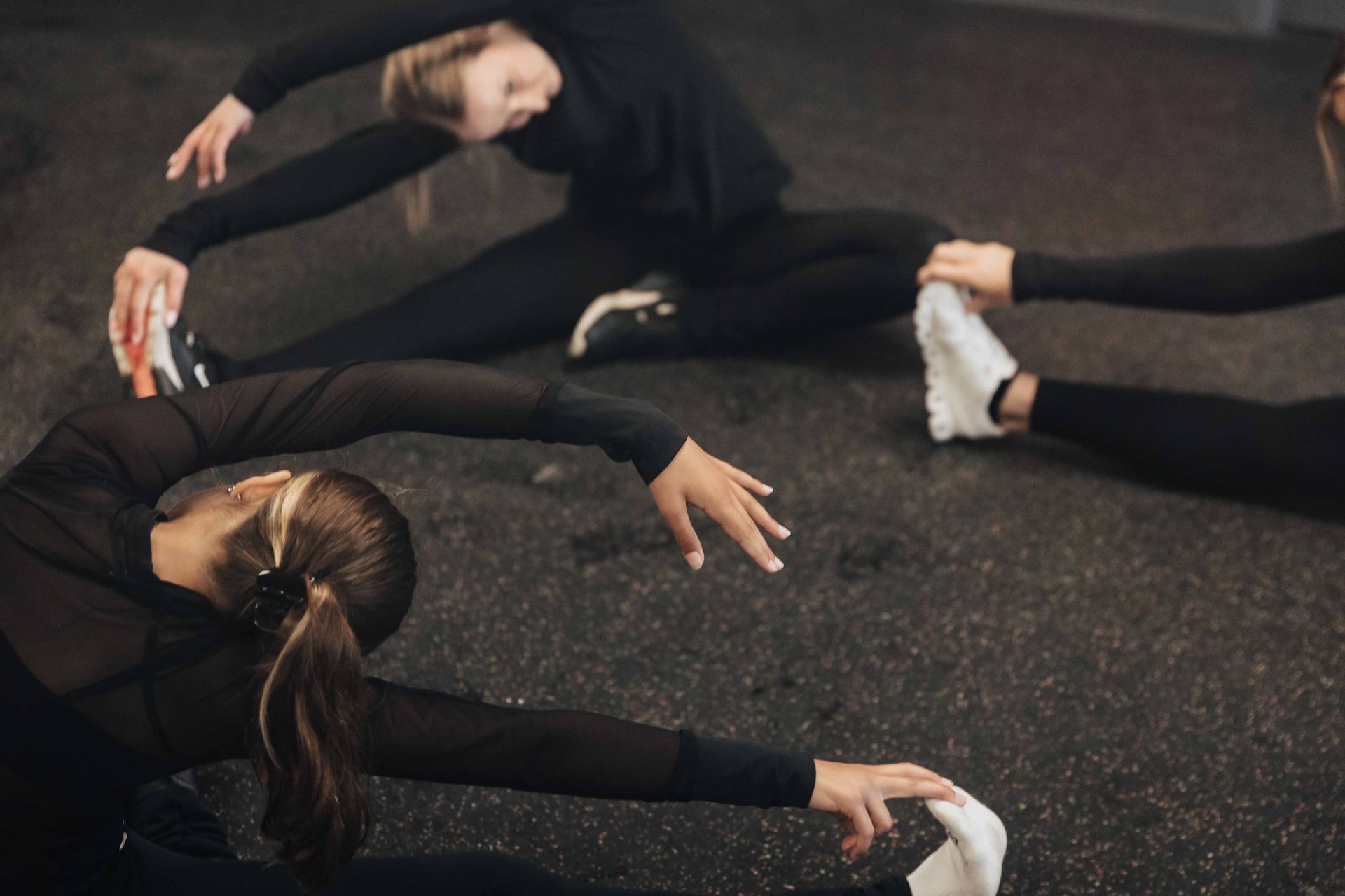 Three people in black activewear stretching on a dark floor, reaching for their feet.