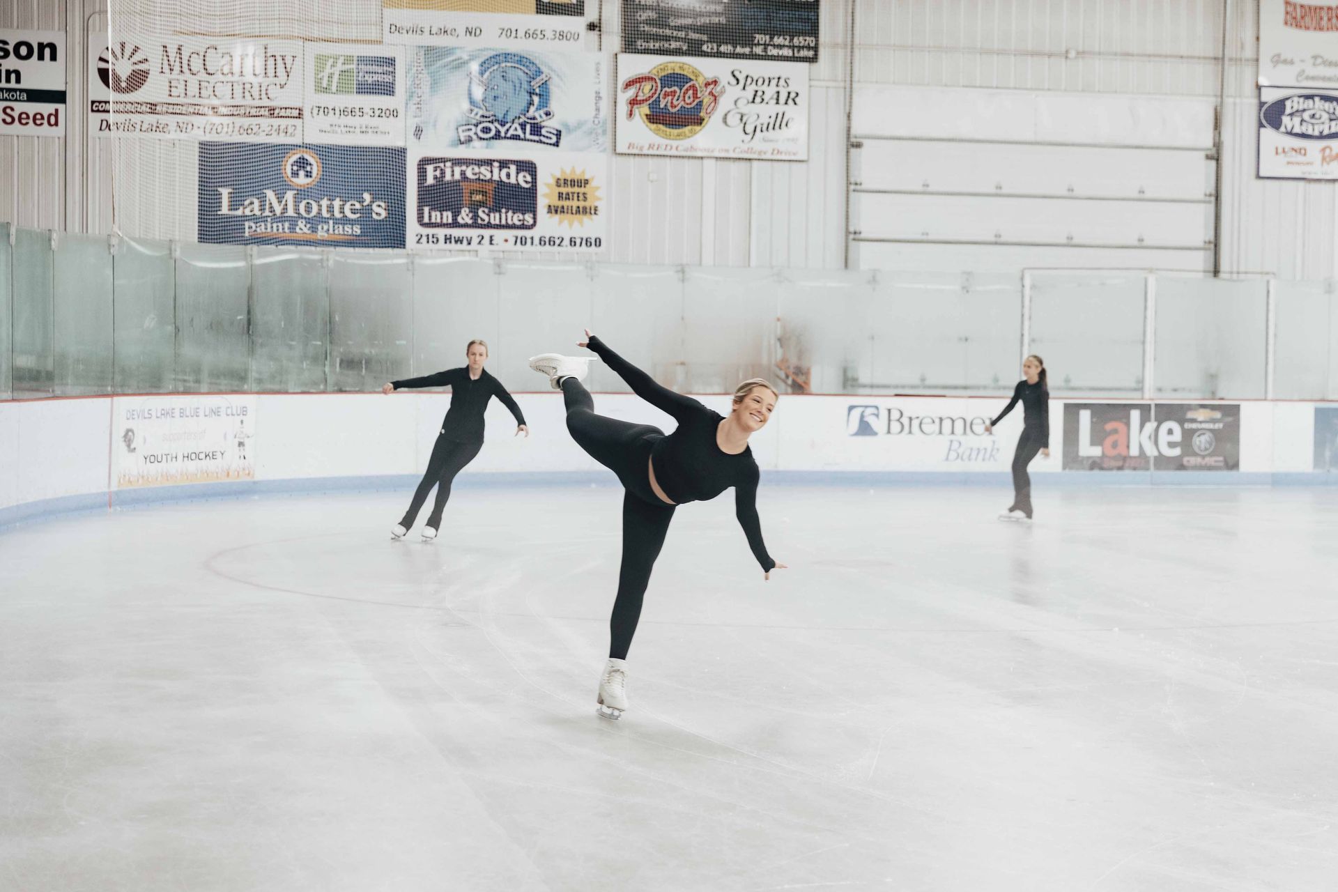 Three ice skaters performing on a rink. One is in a spin; all wear black. The setting is indoors, with signs on the wall.