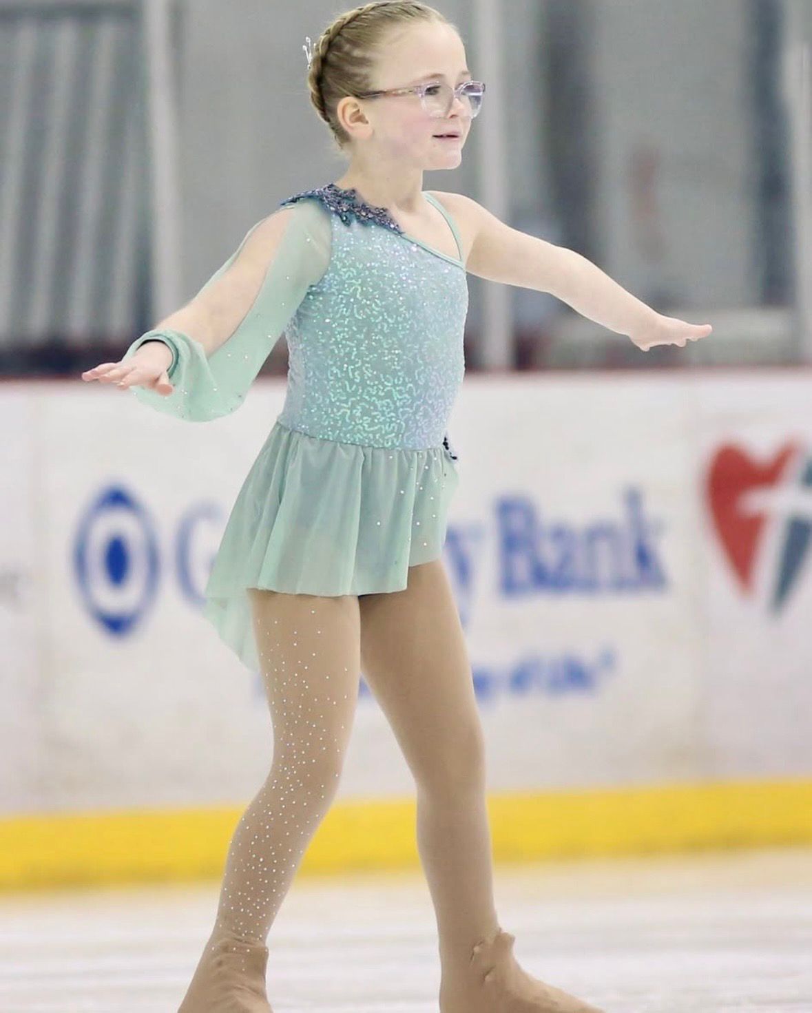Young skater in blue dress performing on ice, arms outstretched.