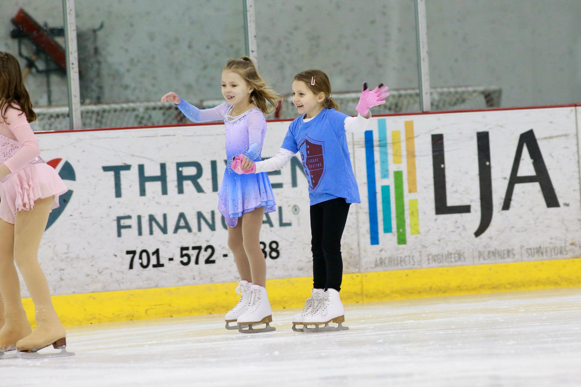 Two young skaters in blue and purple outfits holding hands on an indoor ice rink