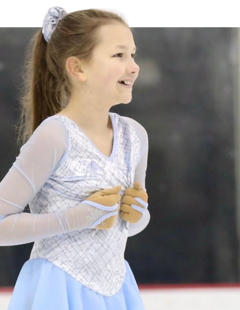 Young figure skater in a pink dress, smiling on the ice rink, with arms extended.